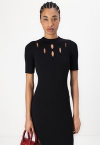 Svart ribbade klänning med korta ärmar och ovala utskärningar längs halsringningen, i kombination med en liten röd handväska. Kroppsnära silhuett.