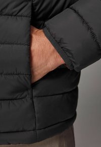 Jachetă puffer neagră cu un model matlasat. Are mânecile cu manșetă și un buzunar lateral, evidențiind un material neted, rezistent la apă.