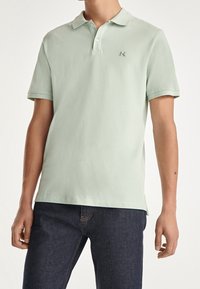 Ljusgrön polo tröja i bomull. Har en krage, placket med två knappar och en liten broderad logotyp på vänster bröst. Kortärmad.