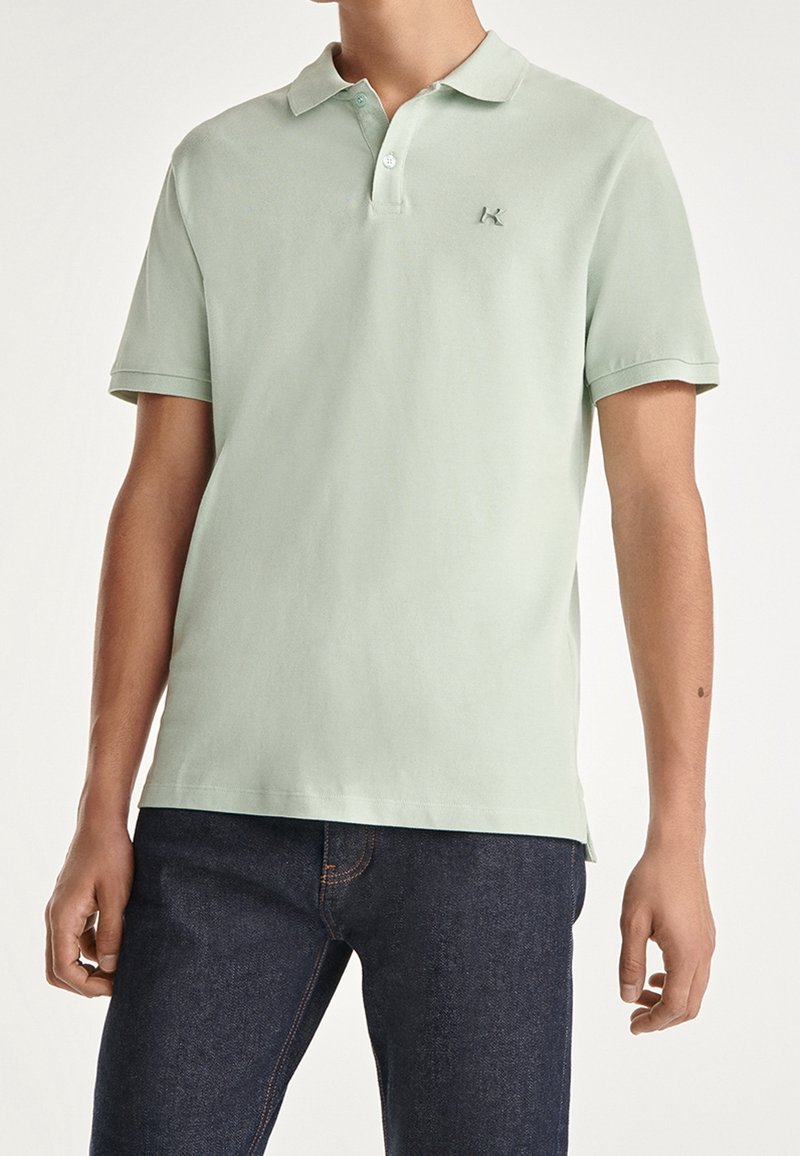 Ljusgrön polo tröja i bomull. Har en krage, placket med två knappar och en liten broderad logotyp på vänster bröst. Kortärmad.
