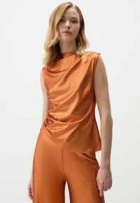 Satin oranje top met een gedrapeerd ontwerp, mouwloos en met een asymmetrische halslijn, gecombineerd met bijpassende high-waisted broek.