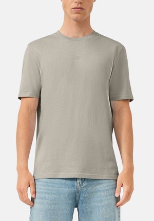 Homme portant un t-shirt à manches courtes gris clair avec un petit logo "QS" sur la poitrine et un jean en denim bleu clair.