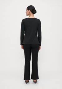 Woman with dark hair in bun wearing black long-sleeve top, black pants, and black heeled sandals, standing facing away against plain background.