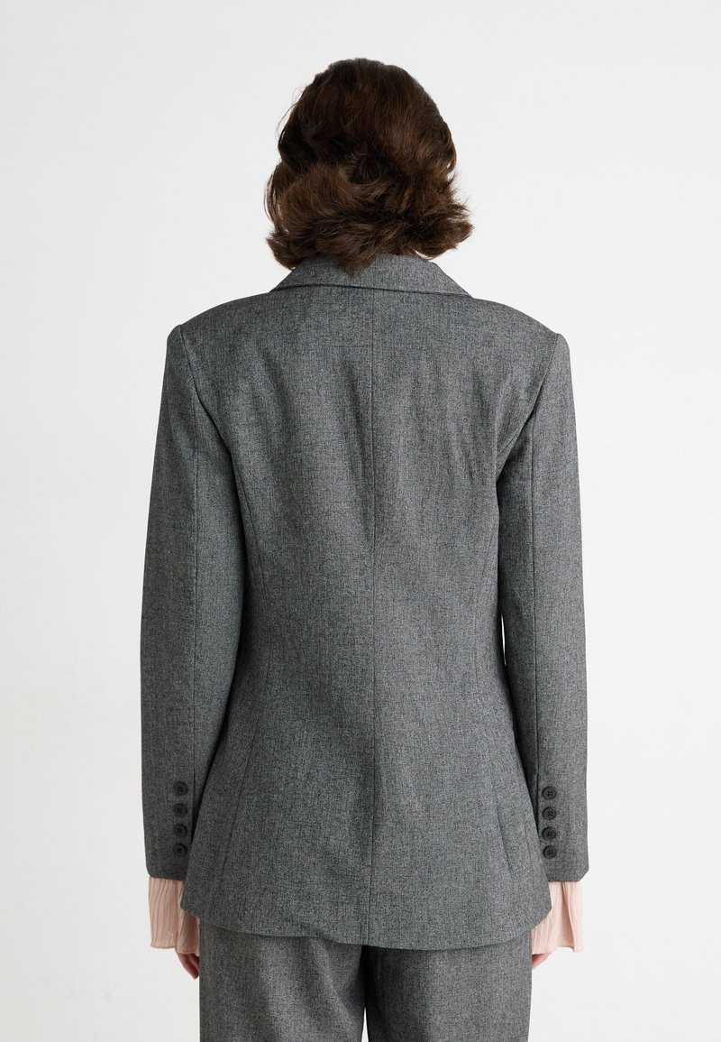 Blazer grigio su misura con vestibilità strutturata, rever a scollatura e polsini a quattro bottoni. Presenta un leggero tessuto testurizzato e uno spacco posteriore singolo.