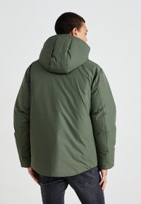 Grønn polstret jakke med hette, med en glatt tekstur og en litt oversized silhuett. Bakbildet fremhever sømmer og detaljene i sømmen.