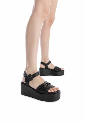 Sandalias de plataforma negras con textura tejida, correa con hebilla y un detalle circular en la parte delantera. Suela de goma con una base gruesa.