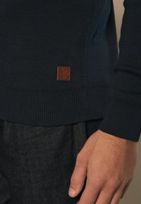 Marineblauwe gebreide trui met geribbelde manchetten en onderkant, met een klein bruin leren merklabel dichtbij de onderkant. G gedragen met donkere jeans.