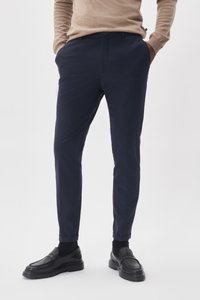 Pantalons navy en tissu à texture lisse, avec un design classique, des poches latérales et des ourlets retroussés. Portés avec des mocassins noirs.