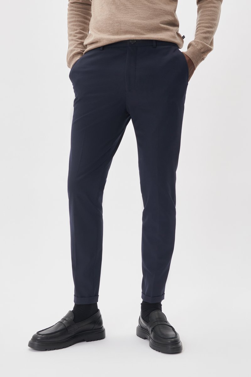 Pantalons navy en tissu à texture lisse, avec un design classique, des poches latérales et des ourlets retroussés. Portés avec des mocassins noirs.