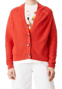 Röd ribbad kofta med v-ringning, knappstängning och strukturerad tyg. I kombination med en vit skjorta och ljusa byxor.