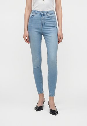Femme portant un jean skinny taille haute bleu clair associé à des talons noirs à bout pointu et un top blanc sans manches, debout devant un fond blanc.