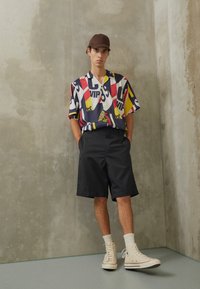 Kortærmet, mønstret skjorte i nuancer af rød, gul, blå og hvid; sorte shorts; beige sneakers; grå betonbaggrund.