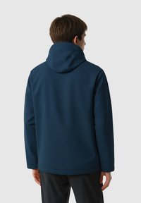 Personne portant une veste à capuche bleu marine, vue de dos, se tenant contre un fond gris clair uni.