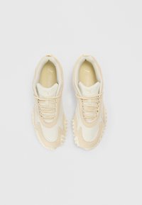 Pantofi sport bej cu partea superioară din plasă, accente texturate și talpă de cauciuc cu pinteni proeminenți pentru aderență. Branț Ortholite vizibil.