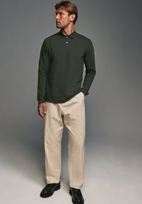 Camisa de polo de manga larga en verde oscuro con un cuello clásico, combinada con pantalones beiges y zapatos negros de cordones, con un corte relajado.