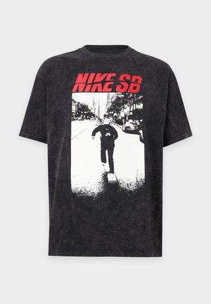 Млад мъж с плитки, облечен в черна тениска Nike SB с червен надпис, черен панталон и бели маратонки, стои пред бял фон.