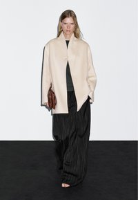 Cappotto di lana beige con un design minimalista, pantaloni a gamba larga a righe nere e borsa in pelle marrone. Modella in piedi su uno sfondo scuro.