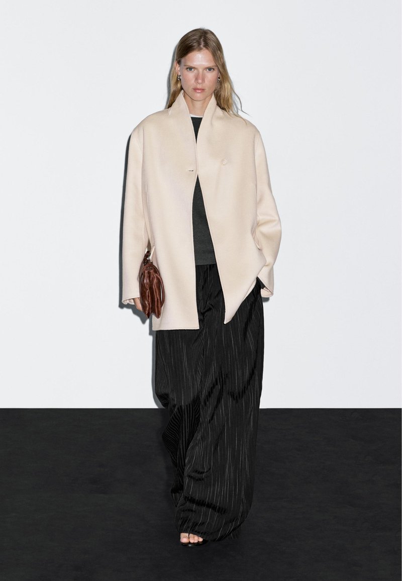 Beige wollen jas met een minimalistisch ontwerp, zwarte gestreepte wijde broek en een bruine leren handtas. Model staat op een donkere achtergrond.