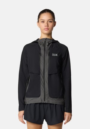 Femme portant une veste de montagne zippée à capuche noire et grise et un short noir, debout devant un fond clair uni.