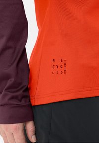Rot und lila langärmliges Shirt mit glatter Textur und sichtbaren Nähten; mit dem bedruckten Text "RECYCLED INSIDE" in der Nähe des Saums.