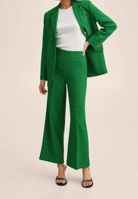 Blazer verde a tinta unita e pantaloni a gamba larga coordinati con tasche frontali, abbinati a una maglietta bianca e sandali neri con cinturini.