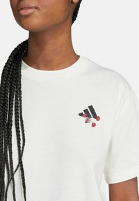 Camiseta de algodón blanca con un gráfico negro que presenta rayas estilizadas y acentos florales rojos en la parte izquierda del pecho. Mangas cortas, escote redondo.