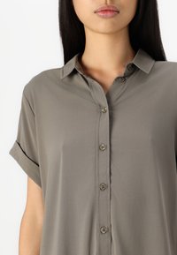 Donna che indossa una camicia grigia a maniche corte, larga e con bottoni, con colletto, in piedi davanti a uno sfondo bianco uniforme.