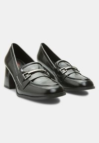 Chaussures à talon bloc en cuir noir arborant un design de mocassin, des coutures marquées et un accent en boucle en métal argenté sur le dessus. Texture lisse.
