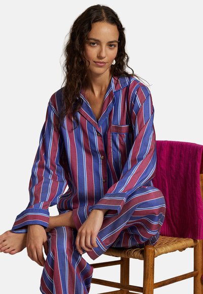 Femme assise jambes croisées sur une chaise en bois, portant un pyjama rayé bleu et bordeaux avec une couverture en tricot rose drapée sur le dossier de la chaise.