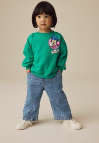 Grøn sweatshirt med et rundt Minnie Mouse grafik og "HEY" tekst. Sammen med blå denim wide-leg bukser og hvide sneakers.