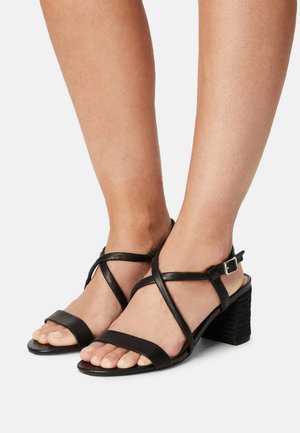 Sorte lædersandaler med krydsede remme og en sort blokhæl. Har ankelrem med sølvspænde. Åben tå-design.