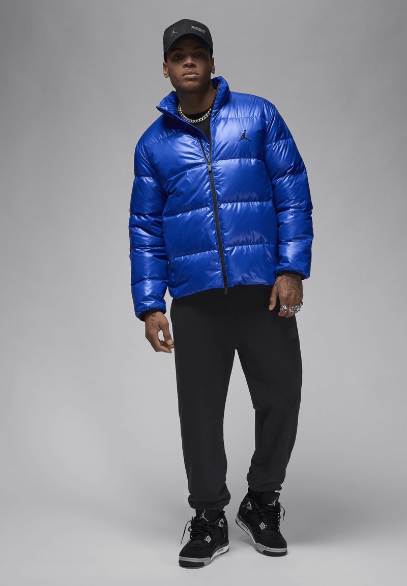 Chaqueta de plumas azul con un acabado brillante, detalles en negro y cierre de cremallera, combinada con pantalones negros y zapatillas con detalles de logotipo.