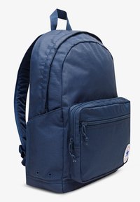 Sac à dos en tissu bleu marine avec une poche avant zippée, des bretelles rembourrées et un patch logo circulaire sur le côté. Matériau texturé et durable.