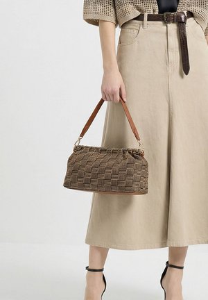 Donna che tiene una borsa intrecciata marrone con manici in pelle, indossa una gonna beige, una cintura marrone e tacchi neri con cinturino alla caviglia.