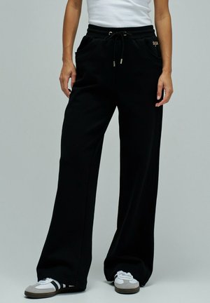 Persona vistiendo pantalones negros de pierna ancha con cintura ajustable y zapatillas blancas, de pie frente a un fondo gris liso.