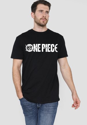 Man wearing black T-shirt with white "ONE PIECE" text and skull with pirate hat logo, paired with blue jeans, standing against gray background.