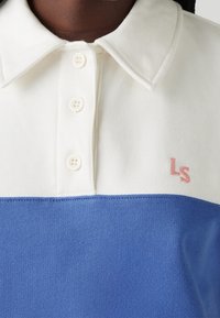 Polo blu e bianco con colletto a punta, caratterizzato da quattro bottoni bianchi e un logo ricamato "LS" rosa in basso a sinistra.