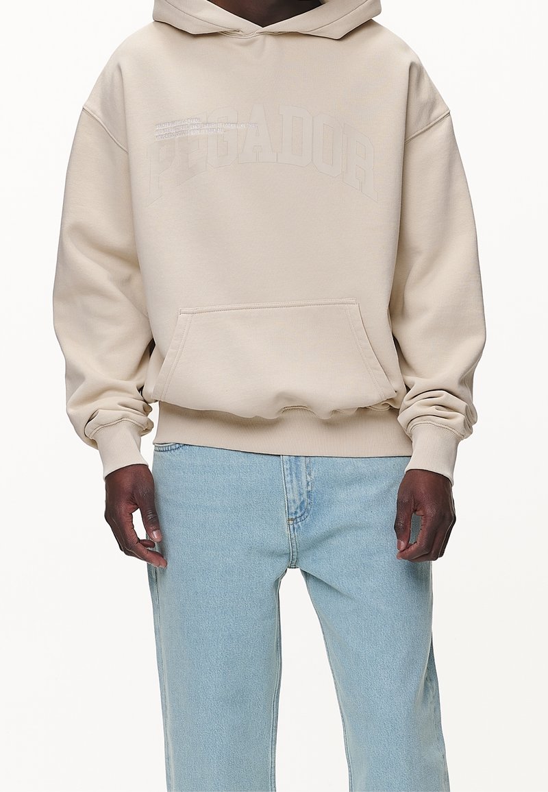 Sweat-shirt beige à capuche avec une poche frontale, des poignets côtelés et un logo en relief. Associe avec un jean bleu clair. Matériel en coton.