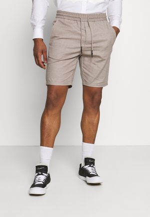 Mann in beigen Shorts mit Kordelzug, weißem Langarmhemd, weißen Crew-Socken und schwarz-weißen Nike-Sneakern steht vor einem weißen Hintergrund.