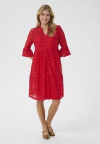 Red lace dress with a V-neck, three-quarter bell sleeves, and a tiered skirt featuring circular cut-out patterns. Brown flat shoes.