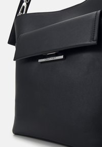 Sac bandoulière en cuir noir Calvin Klein avec fermeture à rabat, fermoir logo argenté et détails métalliques.