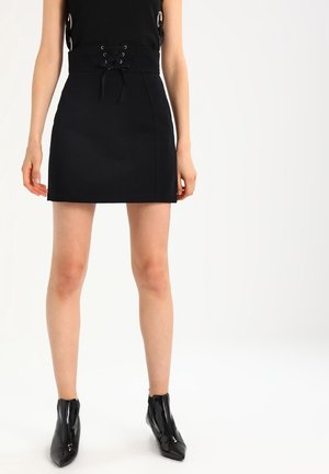 Frau trägt einen figurbetonten schwarzen Minirock mit Schnürungsdetail an der Taille und schwarze Lack-Stiefeletten, stehend vor weißem Hintergrund.