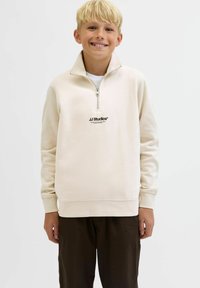 Beige zip-up sweatshirt med hög krage, med "JJ Studios" logotyp i svart och ribbad mudd. Bärs över mörkbruna byxor.