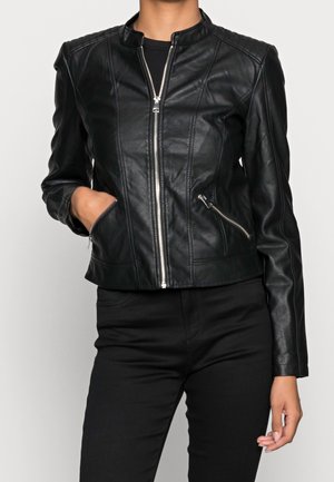 Person trägt eine eng anliegende schwarze Lederjacke mit Frontreißverschluss und Reißverschlusstaschen, kombiniert mit schwarzen Hosen vor einem schlichten Hintergrund.