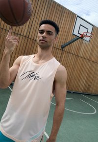 Basketballspieler in einem hellrosa Mesh-Top mit Logo, der einen Basketball auf einem Finger dreht; Holzboden im Hintergrund mit Korb.