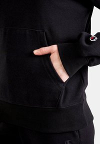 Black sweatshirt with ribbed cuffs and hem, featuring a front kangaroo pocket and a small embroidered logo on the sleeve.