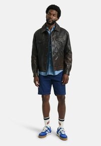 Bruine leren jas met een kraag, blauwe spijkerblouse eronder, marineblauwe shorts en blauwe sneakers met witte zolen en gestreepte sokken.