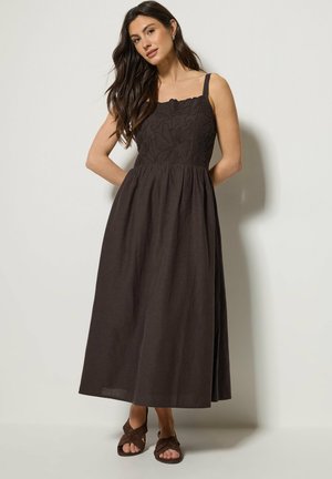 Femme aux cheveux longs et foncés portant une robe marron foncé sans manches avec une broderie florale sur le corsage et des sandales marron à bout ouvert.