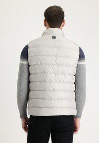 Chaleco acolchado gris claro con textura acolchada, cuello alto y logo circular oscuro en la parte trasera. Mangas de punto en contraste en azul y gris.