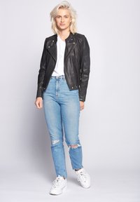 Sort læder bikerjakke med asymmetrisk lynlås, parret med lyseblå, ripped jeans og hvide sneakers. Modellen står foran en lys baggrund.